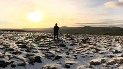 Will Blissett Will standing in a large field, which has snow on the ground, looking out at green hills in the distance. 