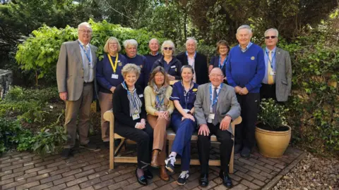 The image shows a group of older adults gathered outdoors in a garden or landscaped courtyard. The scene appears to be taken during daytime, with natural light and leafy greenery suggesting mild weather. The group is posed for a formal or semi-formal group photograph.