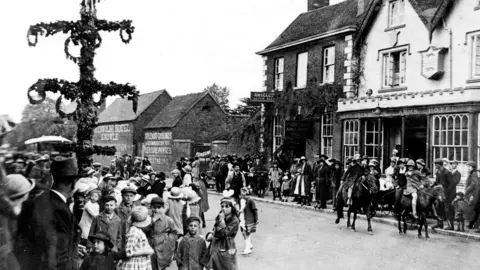 A black and white image showing people gathered in a village high street in the early 20th century. 