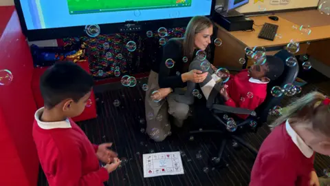 Children and an adult engage in a bubble‑filled activity area in a classroom. A child stands in the foreground wearing a red school jumper, while another child sits in a swivel chair near an adult who is seated on the floor beside them. A board game‑style activity sheet lies on the carpet. In the background, a large interactive screen displays a colourful image, and desks with computers are visible to the right. Bubbles float throughout the scene.