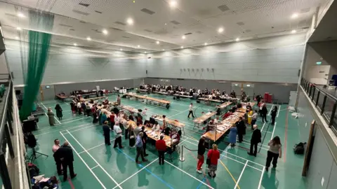 The election count taking place at the Riverside Leisure Centre in Exeter in May 2025 with lines of tables and people sitting and counting votes with people standing around the outside and monitoring the count.
