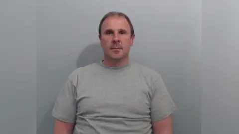 A mug shot of Irvine. It is a head and shoulders image, he is wearing a grey sweatshirt and has short brown hair. 