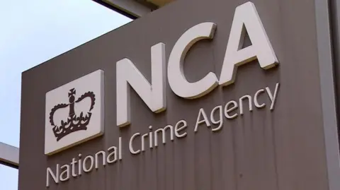 A close up shot of the National Crime Agency headquarters building sign with huge white letters spelling 'NCA' attached to wall. 