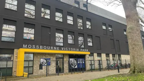 Exterior of Mossbourne Victoria Park Academy, a modern black-and-brick school building with large windows and a yellow entrance canopy, viewed from behind railings with a tree in the foreground.