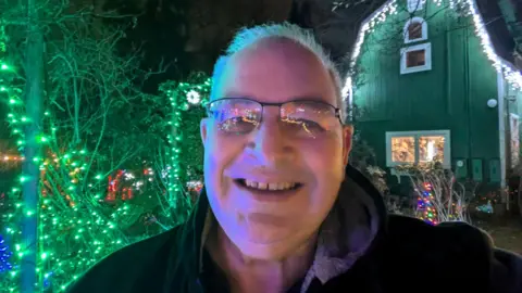 Rik Freeman A selfie of an older man with grey hair and clear-framed glasses. He is wearing a black coat and is standing in front of a house that is decorated with Christmas lights. 