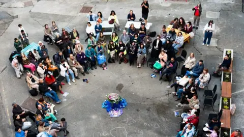 Urmila Doraswami An aerial photo looking down on a circle of people in a courtyard sitting together. There are about 40 people gathered together. 
