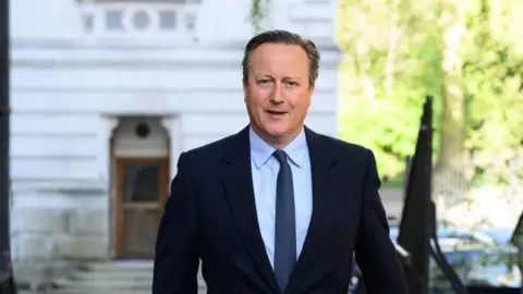 Getty Images The former Prime Minister David Cameron is walking down a street looking at the camera, wearing a navy shirt, light blue shirt and mid blue tie. There is a white building with steps leading up to a brown wooden door in the background and some black railings to the right of the picture.