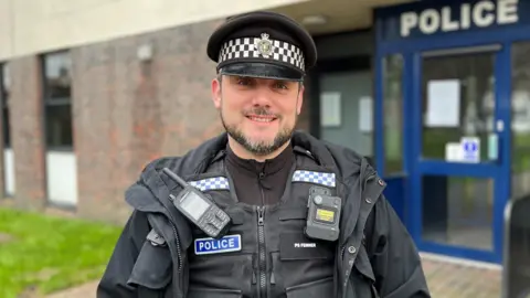 Sophie Seth/BBC A policeman with a dark beard and a police coat and hat on. He is standing in front of a blue door which has "POLICE" written above it in white letters.