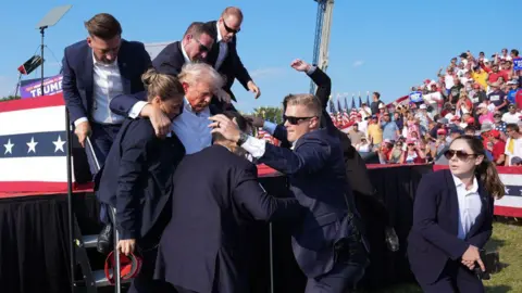 Getty Images Former president Donald Trump is assisted offstage during a campaign rally for former President Donald Trump at Butler Farm Show Inc. on Saturday, July 13, 2024 in Butler, Pa. Trump ducked and was taken offstage after loud noises were heard after he began speaking.