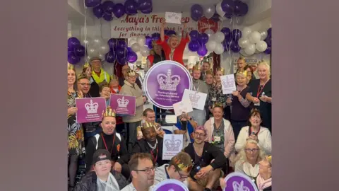Maya's Community Support Centre A group of people holding pictures of a purple crown. There are white and purple balloons in the background.