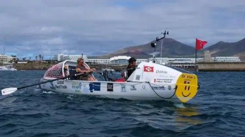 Chloe Foster Tim and Mike sit in their rowing boat on the sea. The hills of Lanzarote can be seen in the distance.