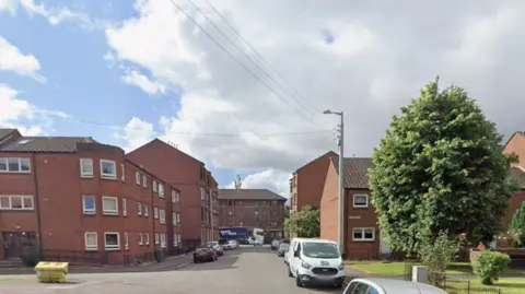 A street view in Glasgow with two and three-storey brick buildings in the foreground and taller ones in the background. There are cars and vans parked on both sides of the street and a large tree in front of the first property on the right. 