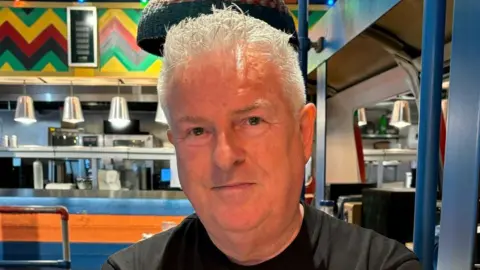 Alex Lawrence smiles at the camera. He has short grey hair and wears a black T-shirt. 