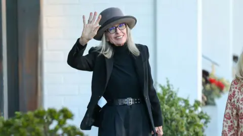 Bellocqimages via Getty Images Diane Keaton photographed on a street in California. She is waving and wearing a turtleneck, thick-rimmed glasses and a hat