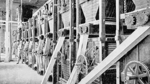 Getty Images A black and white image of a mill with lines of machinery and men looking at the machinery.