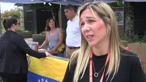 Karolin De Sousa gives a TV interview standing next to a stall to collect aid. She has long, straight, fair hair and wears a black top.