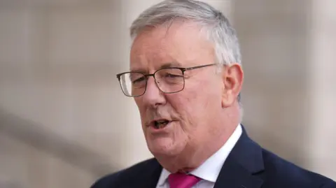 PA Media Mike Nesbitt is wearing a white shirt, pink tie, navy blazer and black framed glasses. He has grey hair and an open mouth. The background is blurry.