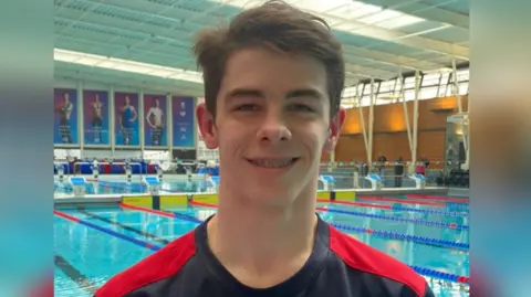 A young man smiles with a swimming pool behind him. 