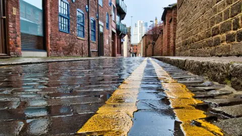 A cobbled road with double yellow lines running down one side. There are buildings on either side of the road. 