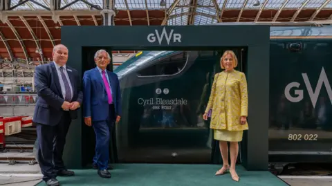 GWR GWR managing director Mark Hopwood and former InterCity managing director Chris Green in navy blue suits with Cyril's widow Katy in a yellow skirt suit on the platform in front of the GWR train named after Cyril Bleasdale OBE.