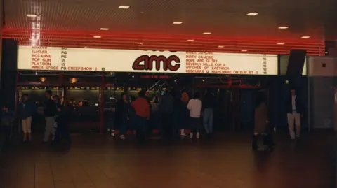 Metrocentre The old cinema which has a lit up board with AMC written in red letters. Some of the films showing are also listed, including Top Gun, Dirty Dancing and Witches of Eastwick. People are gathered below the sign.