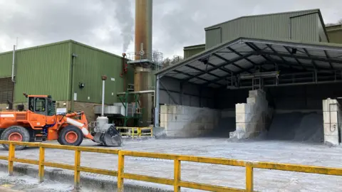 The outside of a factory. On the right of the image, a large storage area with small amounts of grey sand-like material, on the left, an orange front-loader truck in front of a grey factory building with chimney.