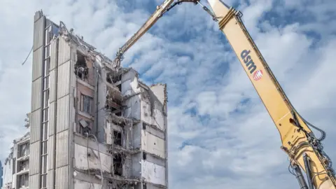 A huge yellow excavator's arm next to a half demolished multi-story building. 