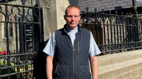 An Anglican rector stands in front of an old church. he is wearing a blue shirt and darker blue jacket. the church building can be seen behind him through iron railings