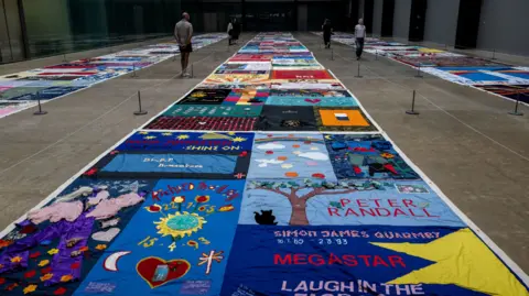 The colourful quilt on show in the Tate. It is in large strips which stretch the length of a large room, and visitors are walking between them.