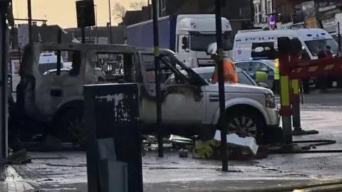 A burnt-out vehicle, with charred bodywork, stands near the scene of the fire and a police van can be seen across the road in Sparkhill.