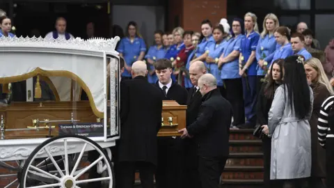 PA Media A white carriage carrying a coffin sits at the bottom of a set of red brick steps. Gathered around it are mourners, some dressed in black and others in the sky and darker blue of the NHS. 