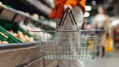 A women holding an empty shopping basket, looking at a supermarket shelf which has fresh veg, including onions, on it