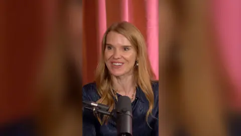 A woman with strawberry blonde hair is sat in front of a red curtain. She's wearing a navy top.