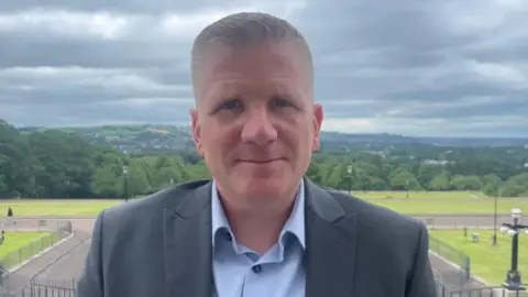 Handout Shows a man with fair hair in a grey jacket and blue shirt with trees and mountains in the background. He is standing on the steps of Stormont.