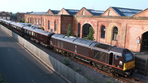 royaltrain.co.uk Five carriages of the royal train are visible on a track, behind it is the old red-brick buildings of Wolverton Works