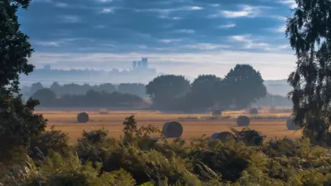 Getty Images An artistic shot of Lincoln city from a distance over misty countryside. There are hay bales in the foreground and Lincoln Minster can be seen silhouetted in the background. 