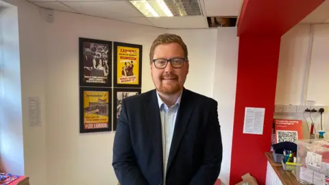 LDRS Head and upper body shot of a man with short reddish hair and a cropped beard, wearing spectacles, a dark jacket and pale blue shirt. Behind him there are four posters displaying old "Vote Labour" adverts