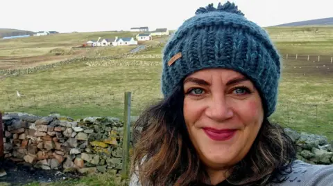 Tammy Hine, smiling and looking towards the camera, wearing a blue knitted hat, she has long dark hair, white houses are behind her, fields and a stone wall. 
