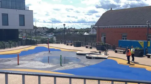 BBC A largely empty paddling pool with a small amount of water in the bed. The floor is electric blue with a yellow border. There are workmen to the right who are wearing hi-vis jackets and are looking at the floor. To the left is a modern building dominated by a glass frontage.