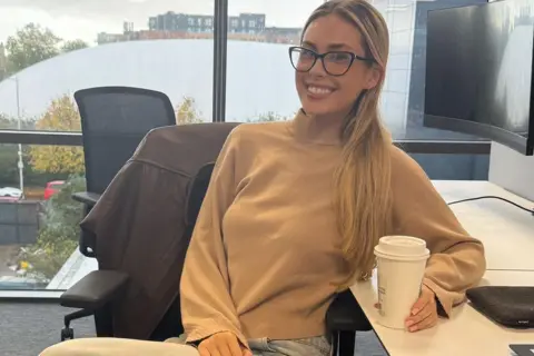 Ellie Jackson A girl smiling in a office setting at her desk holding a coffee cup wearing glasses