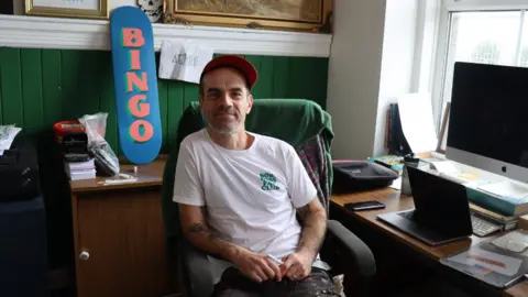 (left image) Ray Bonner in a white t-shirt and red cap, sat at a desk beside a blue graffitied skateboard that says "bingo"