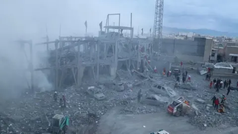 Tasnim People inspect the aftermath of a strike on the Husseinya Mosque. Smoke can be seen rising from the shell of a building while burned out cars are also seen parked outside. 