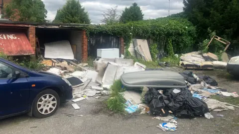 Fly-tipped rubbish next to a row of garages with a dark blue car on the left of the image