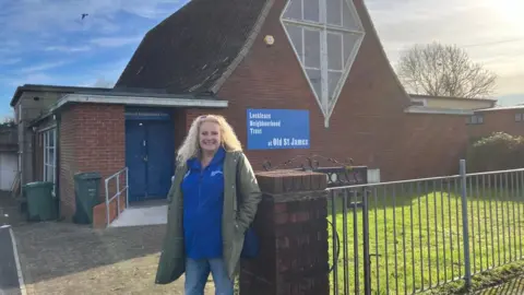 Local activist Maria Perrett poses next to the front gate of the Lockleaze Neighbourhood Trust which is in the old St James Presbitarian Church. It is a brick building with a  blue door and a diamond shaped window on its front.