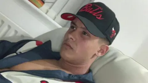 A young man wearing a blue, white and red shirt, open to the chest, and a black baseball cap with "Chicago Bulls" on the front is sitting on a white sofa 