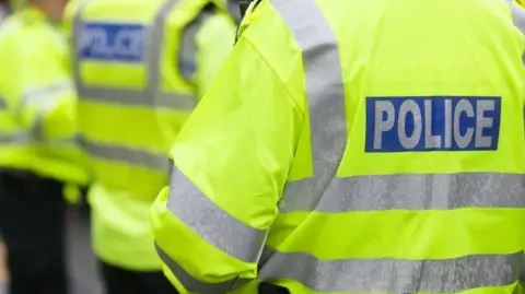 Three police officer are standing next to each other. They are all wearing yellow hi-vis jackets with silver stripes, with police written on the back in silver. 
