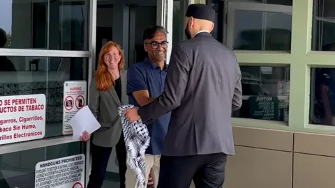 Badar Khan Suri smiling while meeting a man holding a keffiyeh, with a woman standing behind him