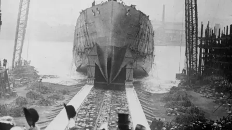 Getty Images A black and white image of the launch of HMS Queen Mary at Palmer's Shipbuilding, Jarrow-on-Tyne. In the foreground black hats are raised to wave off the ship as she rolls down rails and into the River Tyne. 