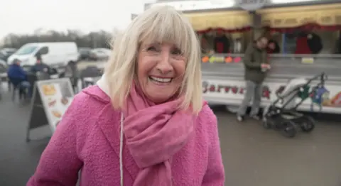 Phillip Norton/BBC A lady with blonde hair smiling at the camera. She is wearing a pink-coloured fleece and matching scarf. There is a food stall in the background.
