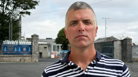 Ed Hill looks to the camera, a head and shoulders shot outside military barracks. He wears a navy blue and white stripe polo shirt.